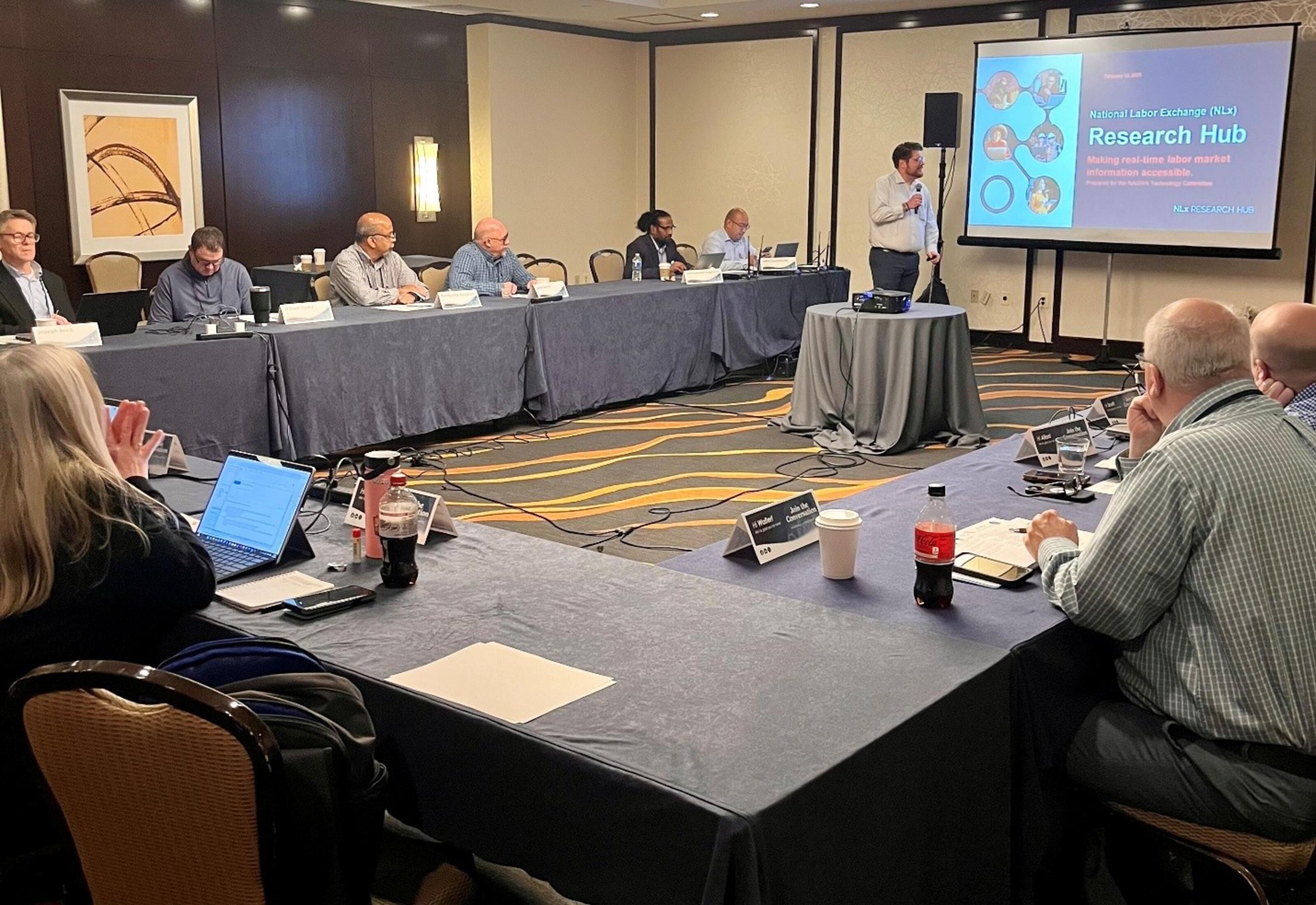 Paul Daniels, NLx Research Hub Manager presents at NASWA's Winter Policy Forum in Washington, DC. Attendees are seated around the presenter who is gesturing toward a screen.